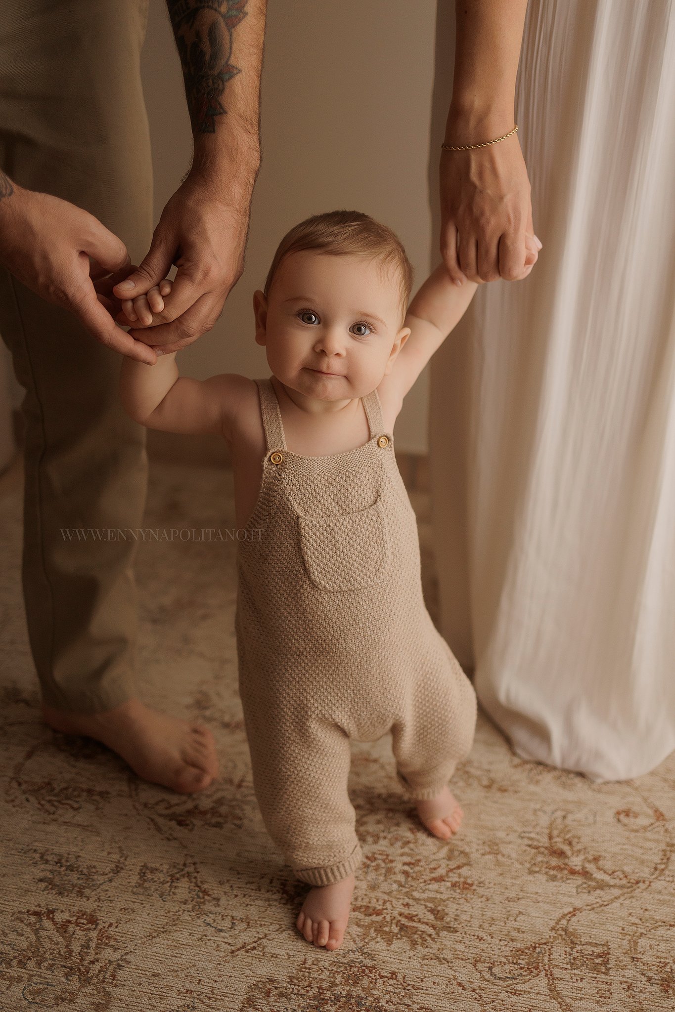 Fotografia di un bambino di 7 mesi che fa i primi passi e viene tenuto dalle manine dai genitori