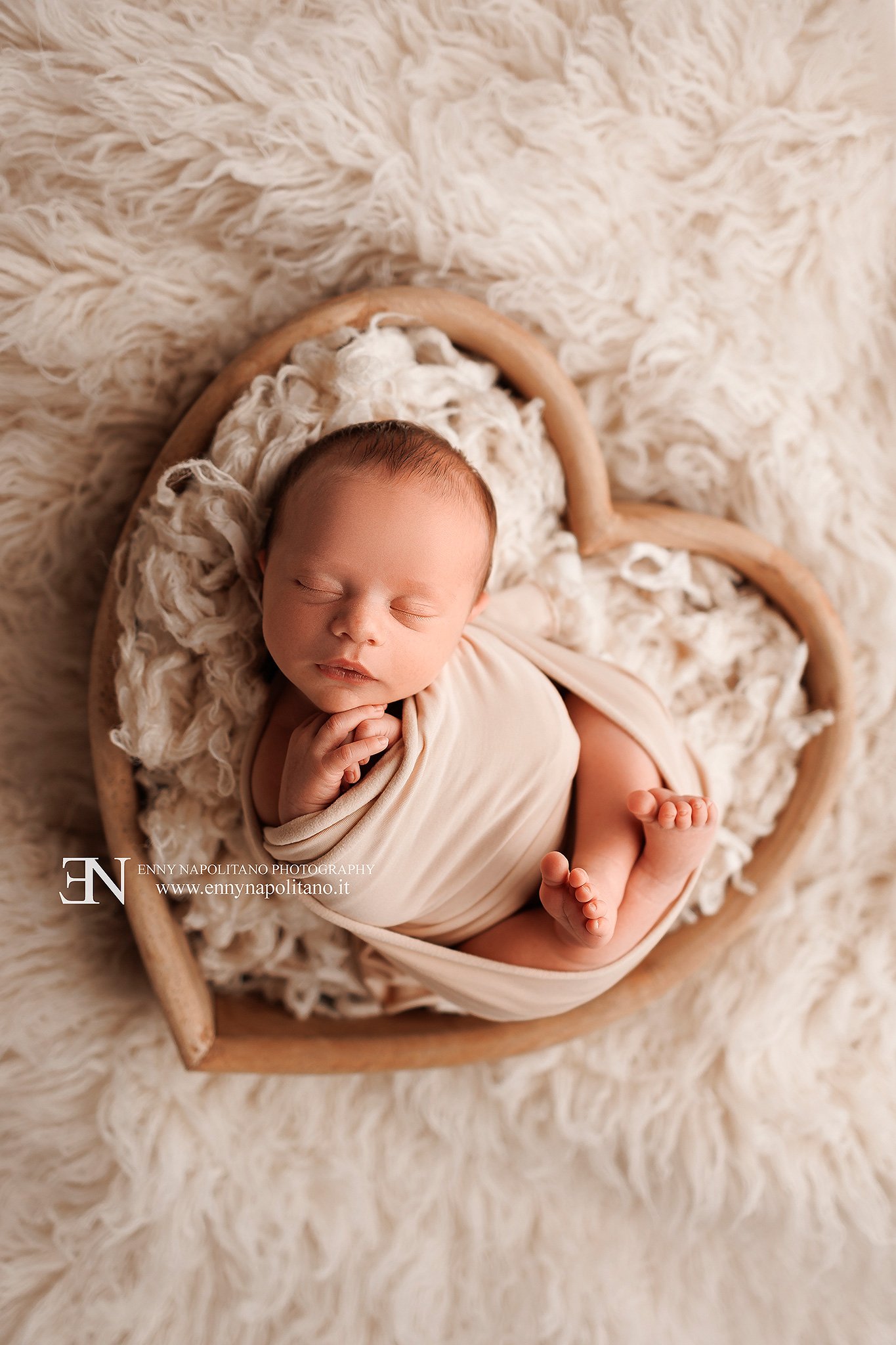 Fotografia newborn di un neonato fasciato che dorme in un prop a forma di cuore in un set color neutro